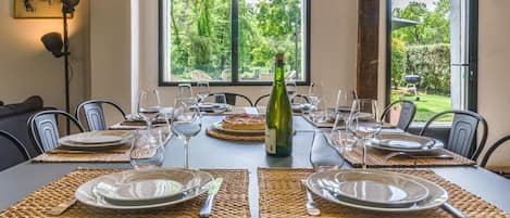 A dining table set for six with placemats, plates, cutlery, and wine glasses. A bottle of wine is placed in the center. The room has a view of greenery through large windows.