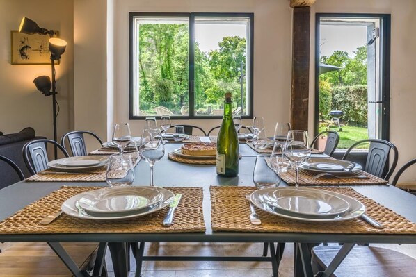 A dining table set for six with placemats, plates, cutlery, and wine glasses. A bottle of wine is placed in the center. The room has a view of greenery through large windows.