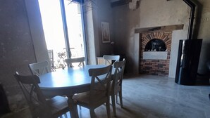Un comedor con una mesa redonda y seis sillas, una lámpara colgante, una chimenea de ladrillo y vigas de madera a la vista en el techo.