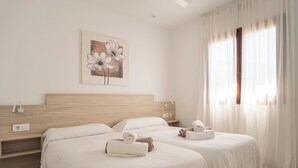 A hotel room with two beds, a wooden headboard, a painting of flowers, and a window with curtains. - V110B | Villa La Pau (Son Bou)