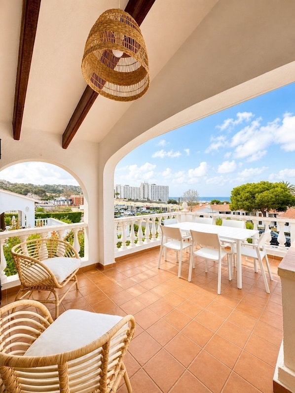 Un balcón con mobiliario blanco, una lámpara colgante de mimbre y vistas a edificios y follaje.