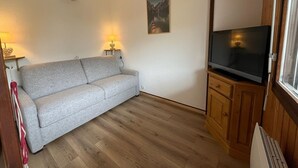 Una sala de estar con un sofá gris, piso de madera, un televisor de pantalla plana sobre un mueble de madera y un cuadro en la pared.