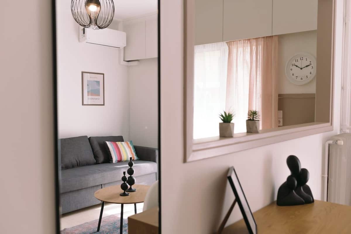 Uma sala de estar moderna com um sofá cinza, uma mesa redonda pequena, um espelho e um relógio na parede.