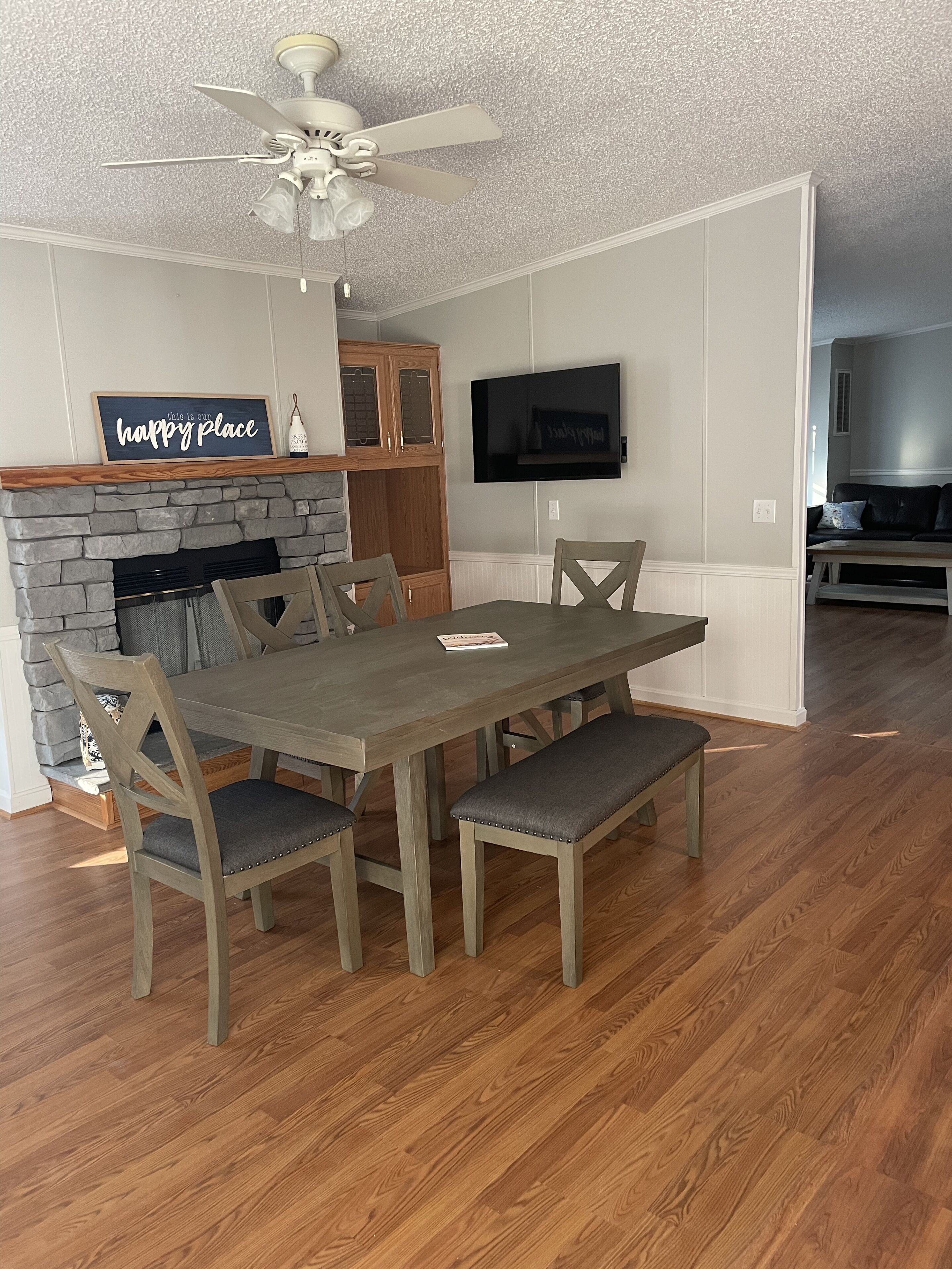 A dining area with a stone fireplace, wooden cabinets, a flat-screen TV, and a ceiling fan.