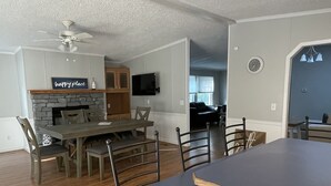 A spacious living room with a large dark-colored dining table, wooden chairs, and a fireplace. A television is mounted on the wall, and there is a ceiling fan. - Minutes to the Beach with a hot tub and decks for relaxing. (Ocean View)