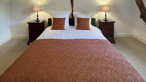 Un dormitorio con techo inclinado, vigas de madera a la vista, una cama grande con ropa de cama blanca y marrón, y dos mesitas de noche con lámparas.