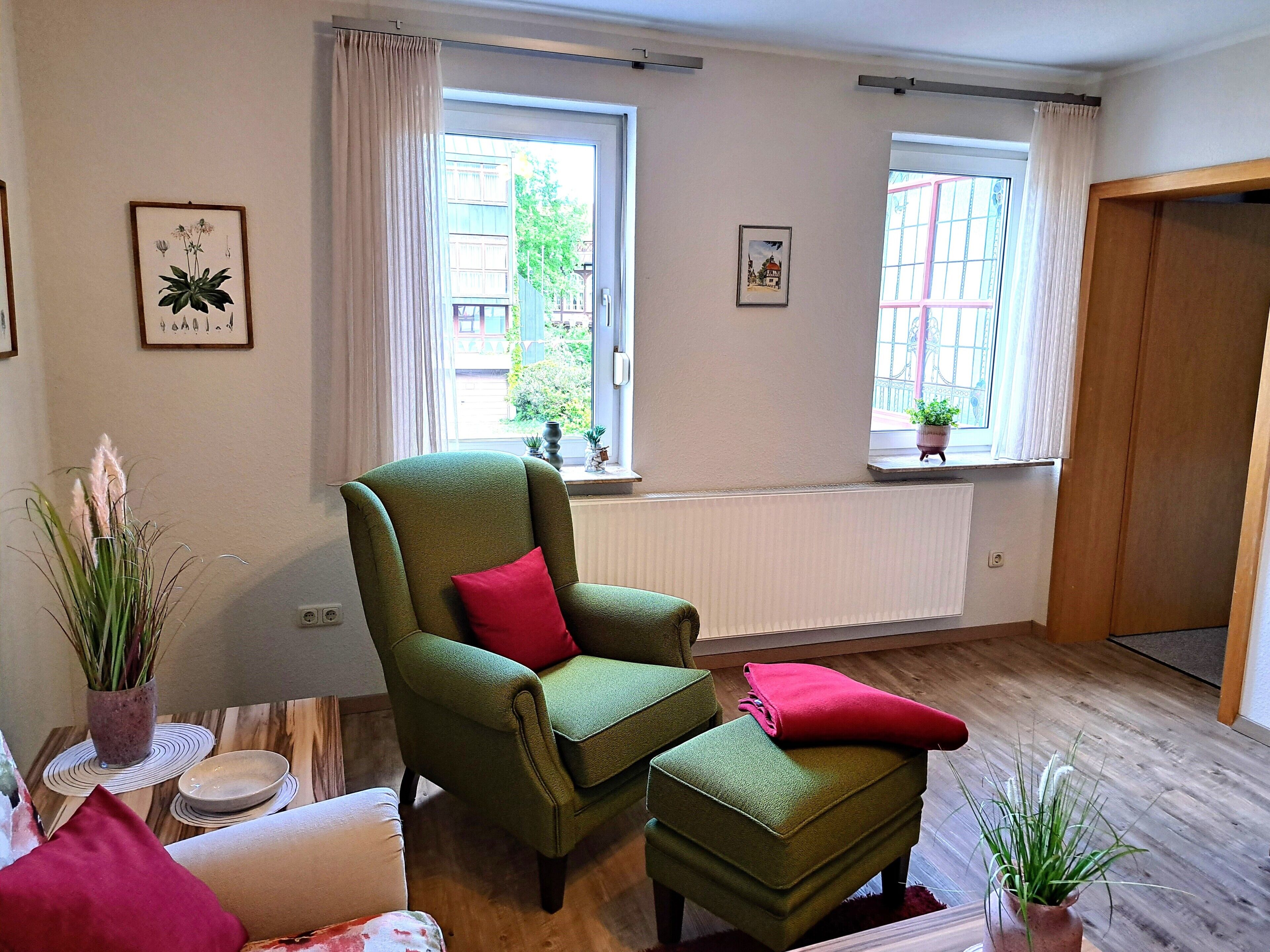 Ein Zimmer mit einem grünen Sessel, dazu passendem Hocker, einem kleinen Tisch mit Schüssel und Teller, und einem Fenster mit Blick auf Gebäude und Grünflächen.