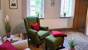 Una habitación con un sillón verde, un reposapiés a juego, una mesita con un bol y un plato, y una ventana con vistas a edificios y zonas verdes.