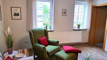 Ein Zimmer mit einem grünen Sessel, dazu passendem Hocker, einem kleinen Tisch mit Schüssel und Teller, und einem Fenster mit Blick auf Gebäude und Grünflächen.