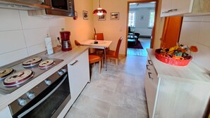 Cocina equipada con estufa, microondas y cafetera. Hay un comedor con mesa y sillas. Se ve una cesta de fruta sobre la encimera.