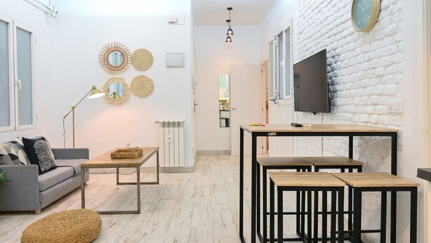 A modern living room with a sofa, coffee table, and ottoman, a dining area with a table and chairs, and a TV mounted on a white brick wall. - Moderno y elegante ap. Barrio Ibiza MEN30 (Madrid)
