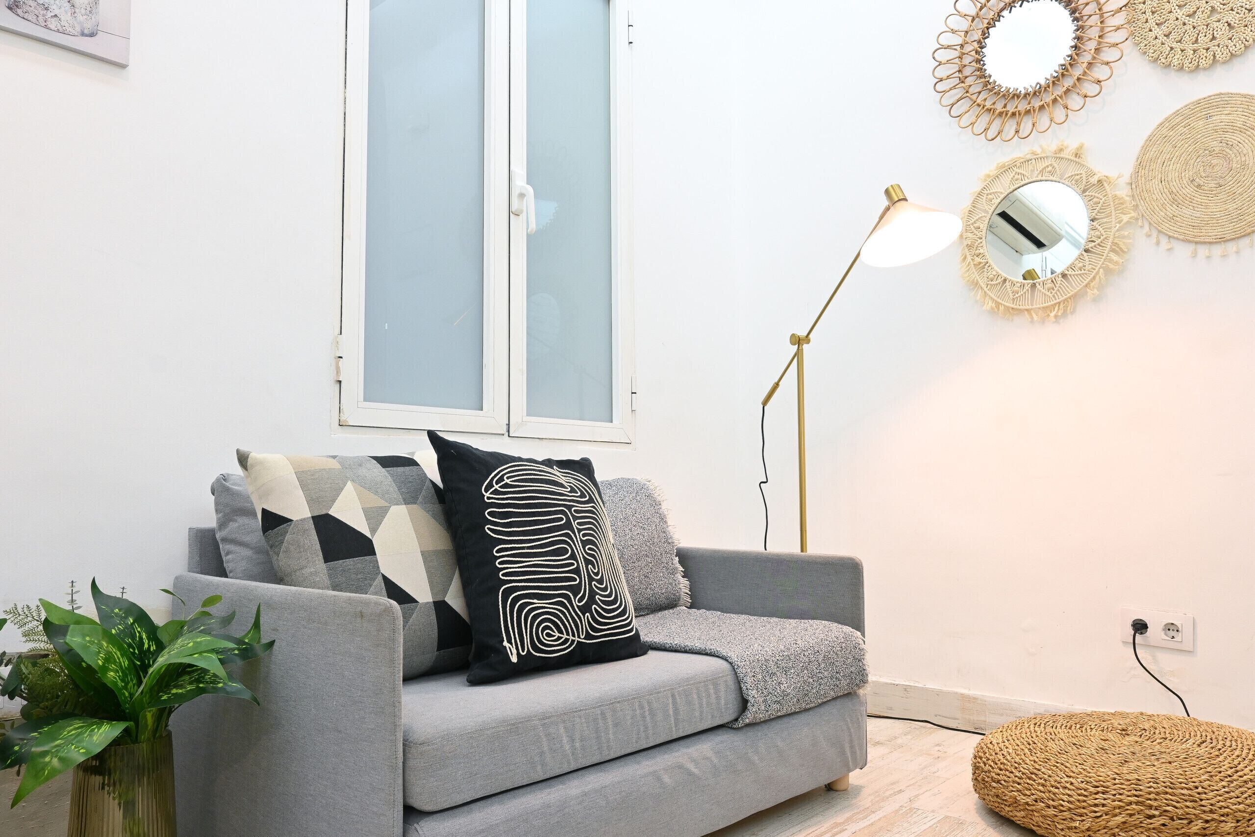 A modern living room with a grey sofa, a black and white patterned pillow, a floor lamp, and a woven pouf.
