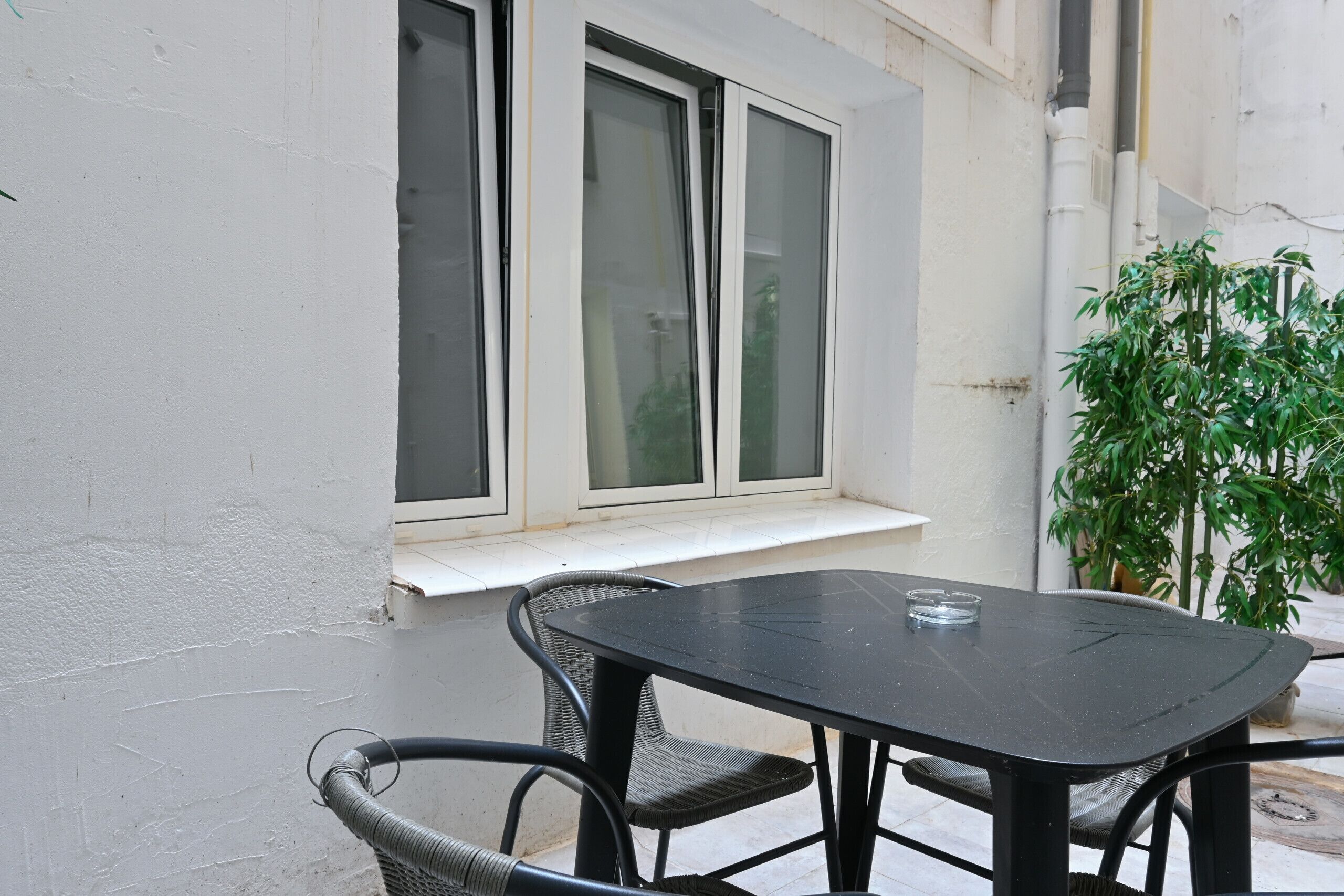 A small outdoor patio with a table and chairs, a window with a white sill, and a potted plant.