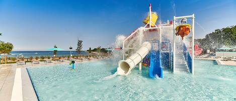 A water play area with a slide and water jets, surrounded by a pool and a view of the ocean.