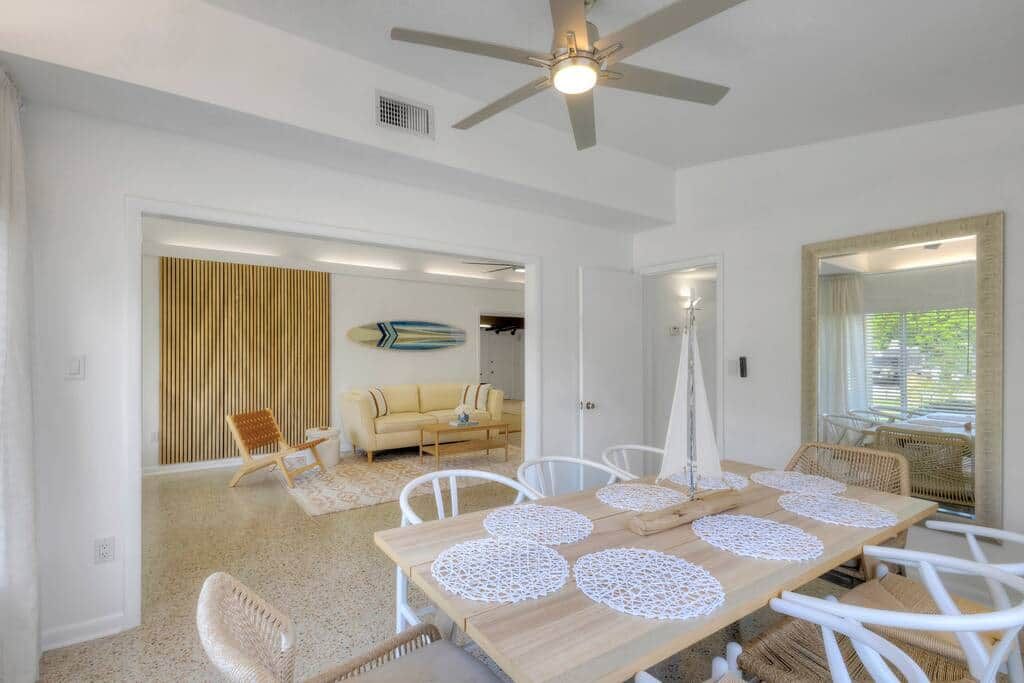 Una habitación amplia con una mesa de comedor de madera, sillas blancas, un ventilador de techo y un espejo grande.