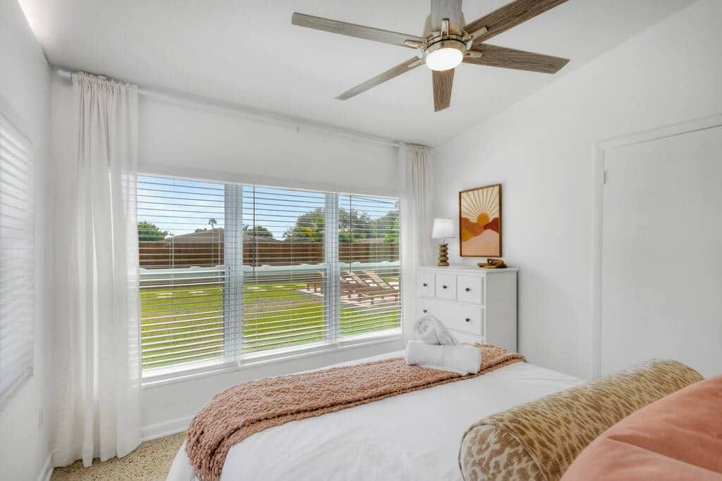 Ein Schlafzimmer mit einem Bett, einem Deckenventilator, einem Fenster mit Jalousien, einem Schminktisch und einem gerahmten Bild an der Wand.