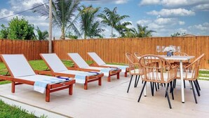 A patio with wooden lounge chairs and a table with chairs, surrounded by a wooden fence and palm trees.