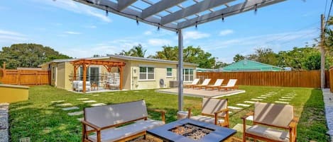 A backyard with a pergola, outdoor seating, a fire pit, and a house in the background.
