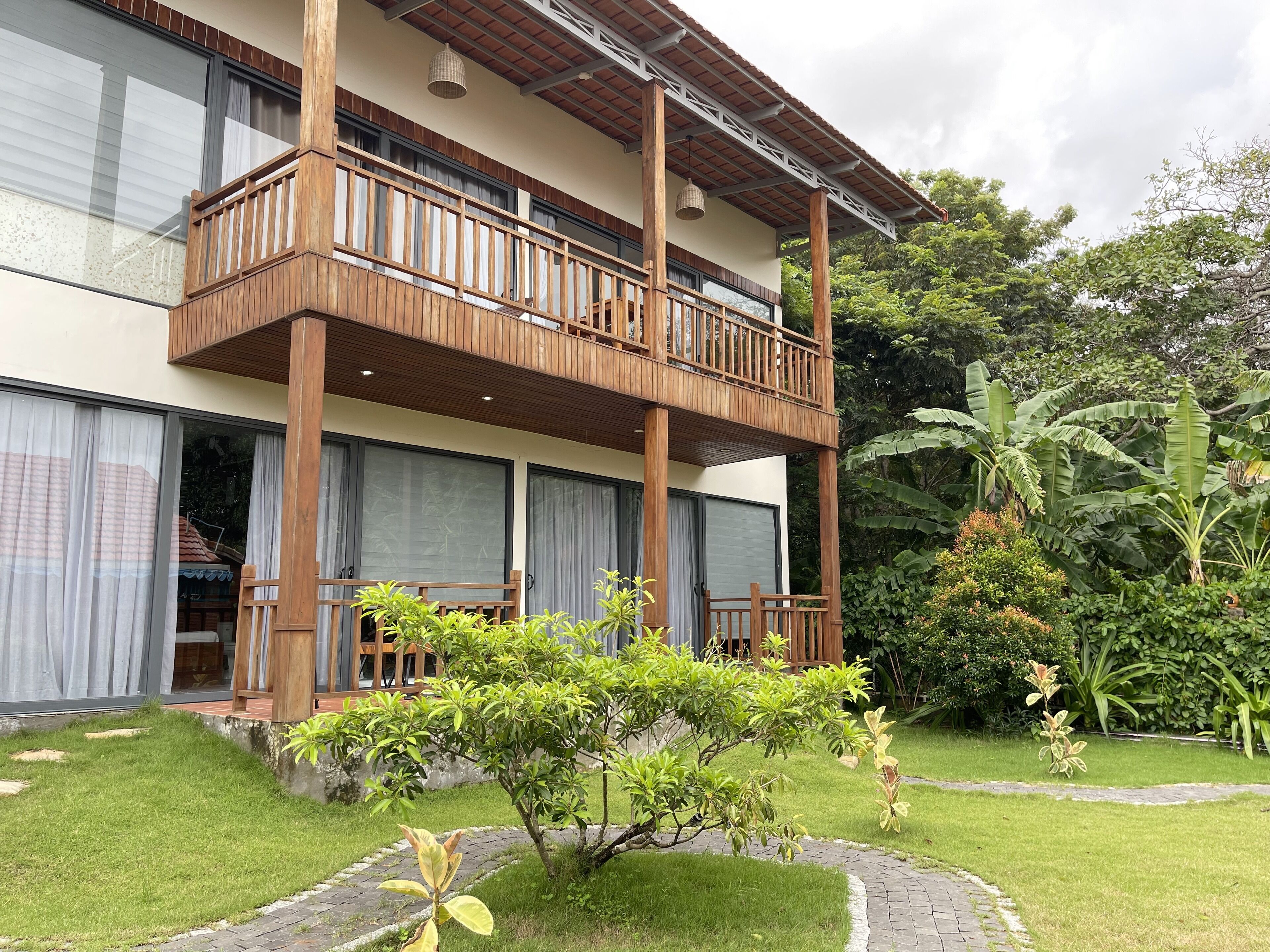 A two-story building with a wooden balcony, large windows, and a garden with various plants.