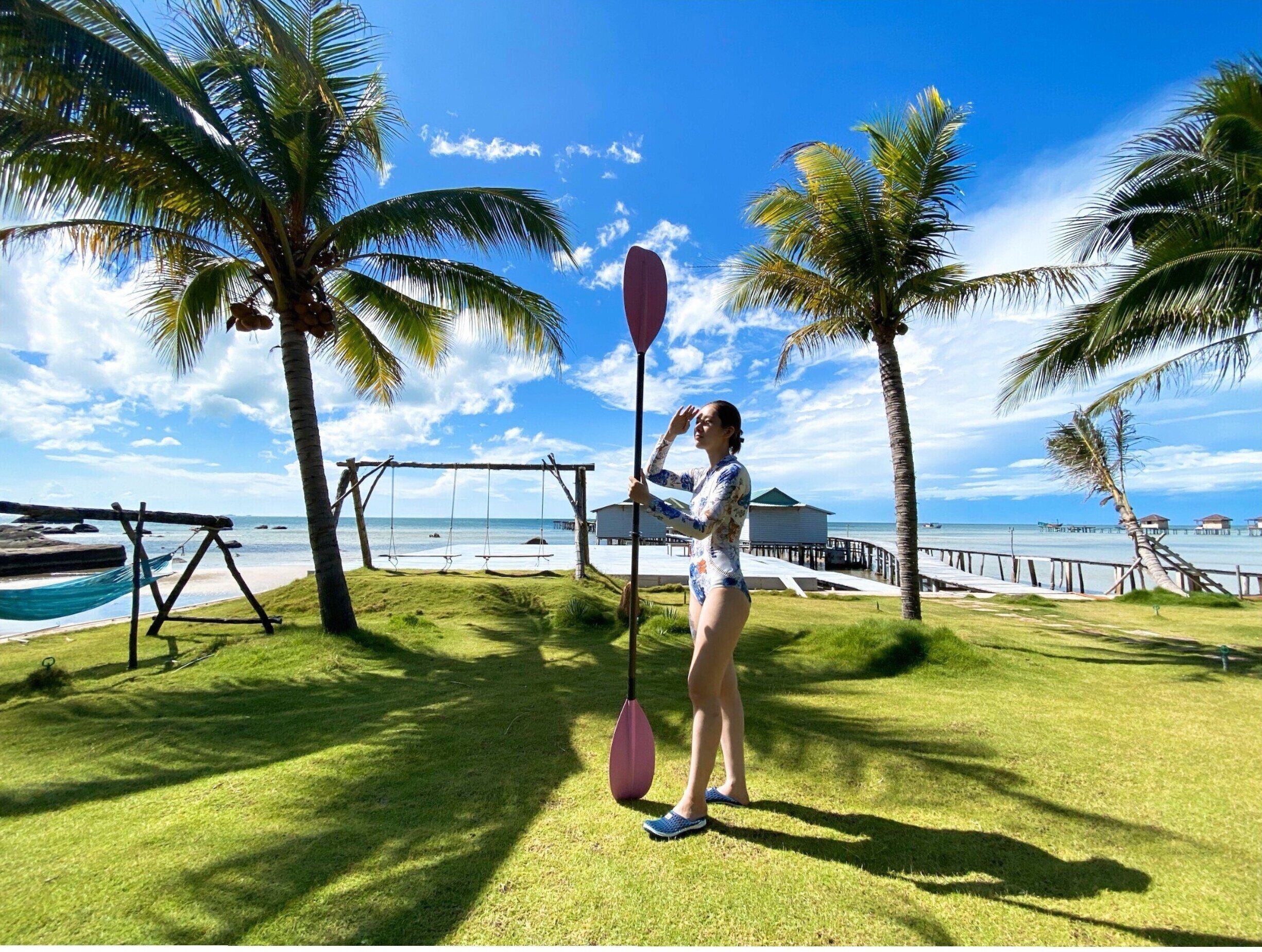 A person standing on a grassy area with a swing set and a hammock, overlooking a beach with palm trees and a clear sky.