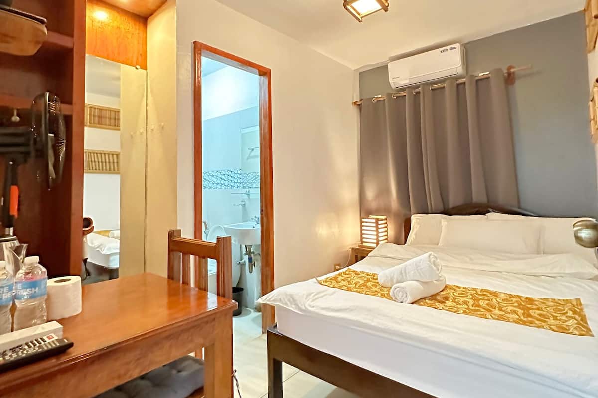 Une chambre d’hôtel avec un lit, un bureau en bois, une chaise, une salle de bain visible à travers une porte ouverte et un appareil de climatisation.