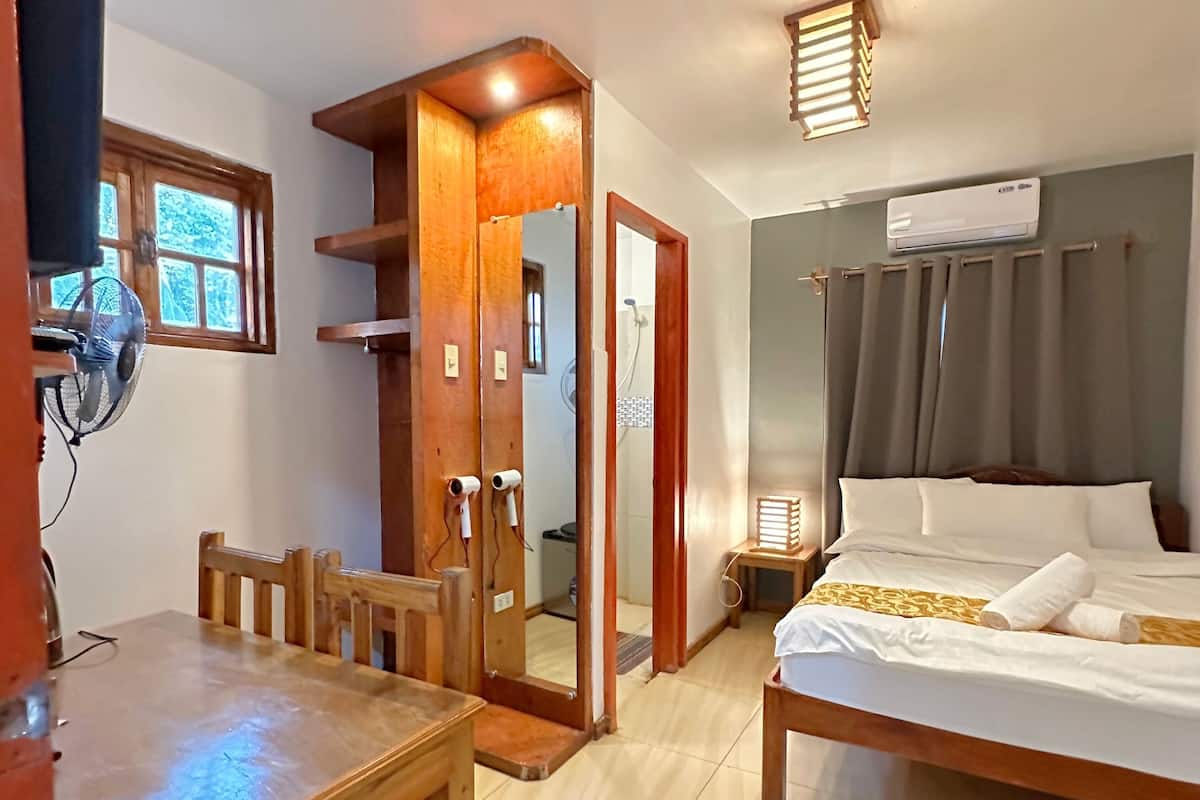 Une chambre d’hôtel avec un lit en bois, une petite table avec des chaises, un miroir et une fenêtre avec des stores.