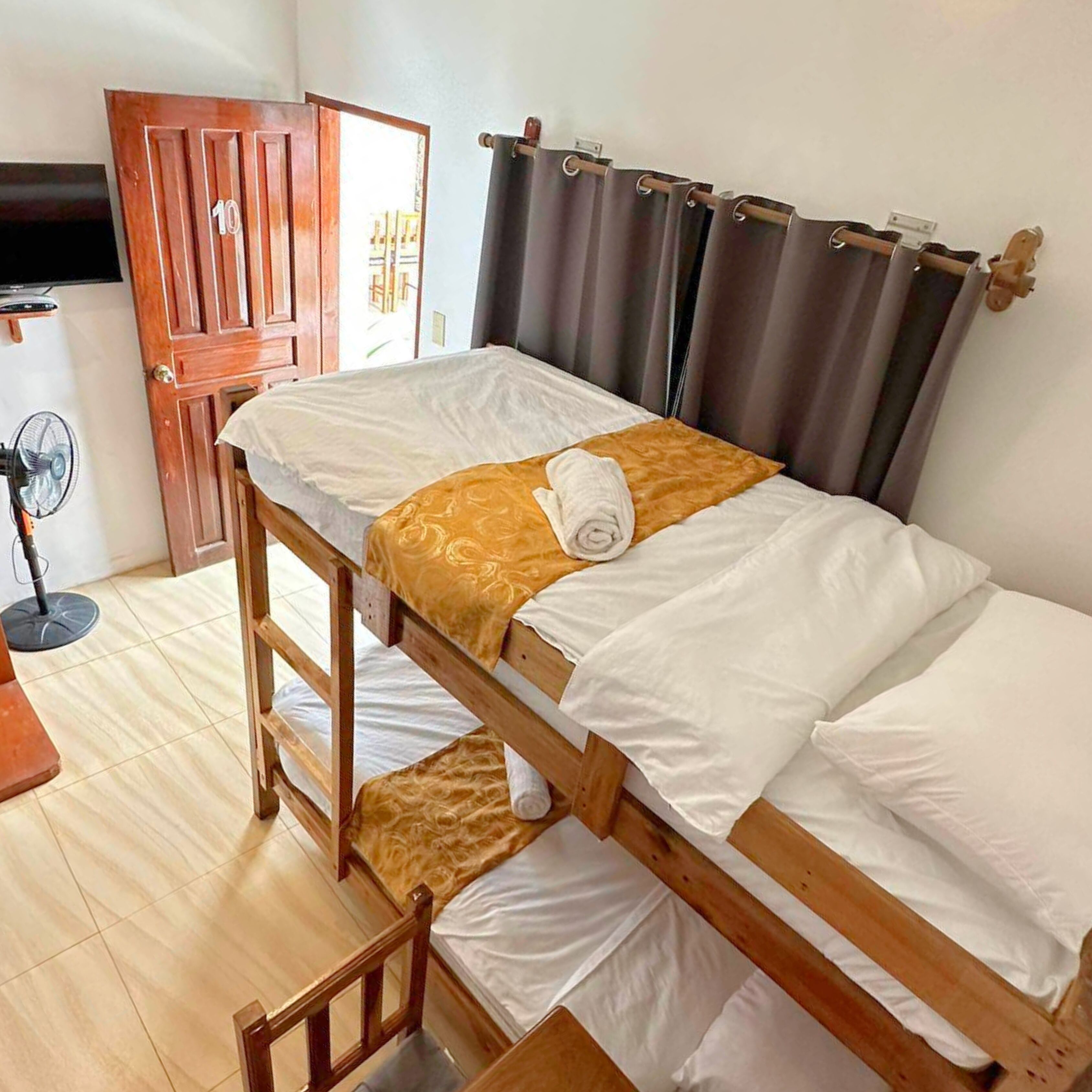 A single-bed room with a wooden frame, white bedding, a towel, a fan, and a door with a number.