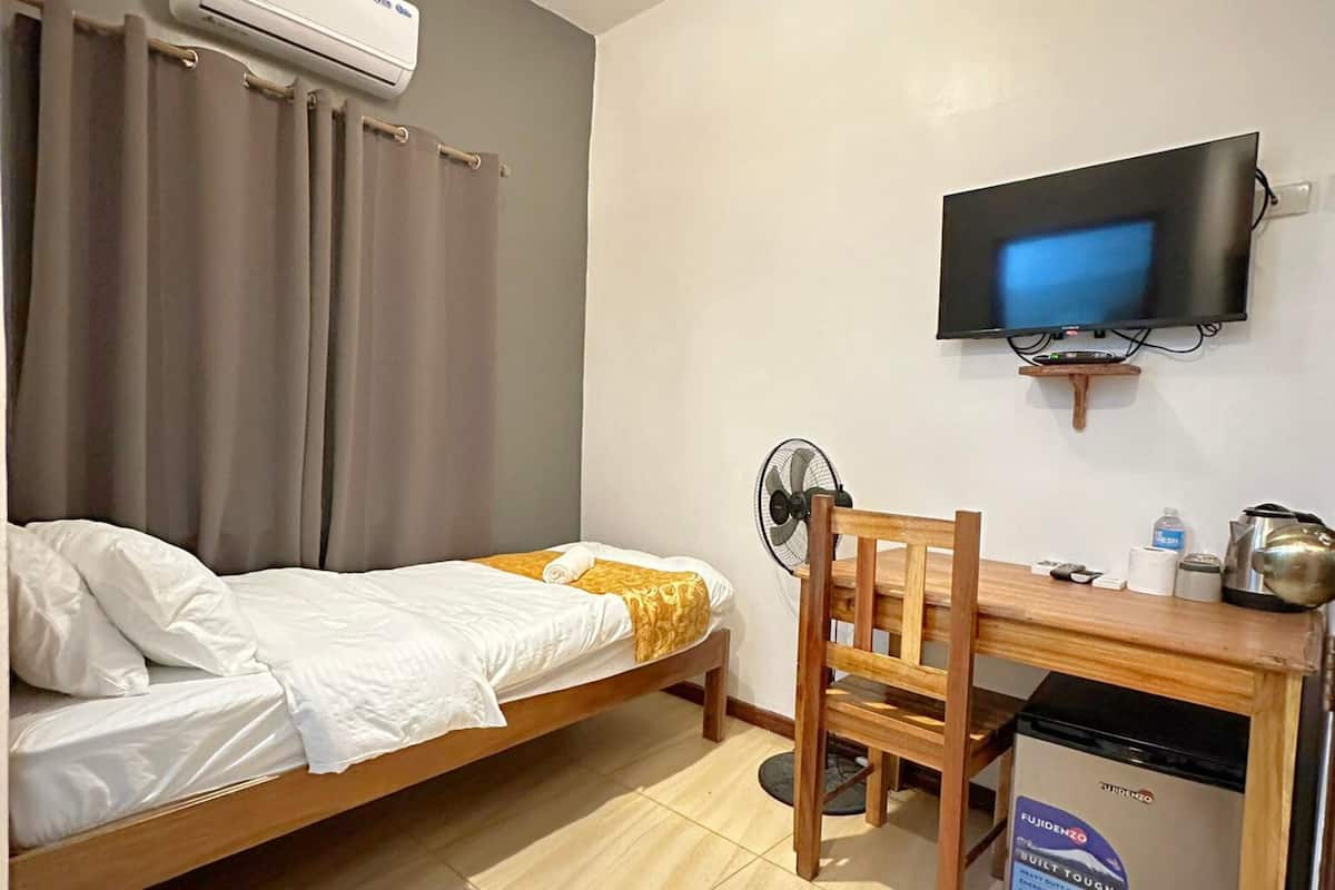 Une chambre d’hôtel de taille réduite, comprenant un lit simple, un bureau avec une chaise, une télévision et une petite kitchenette.