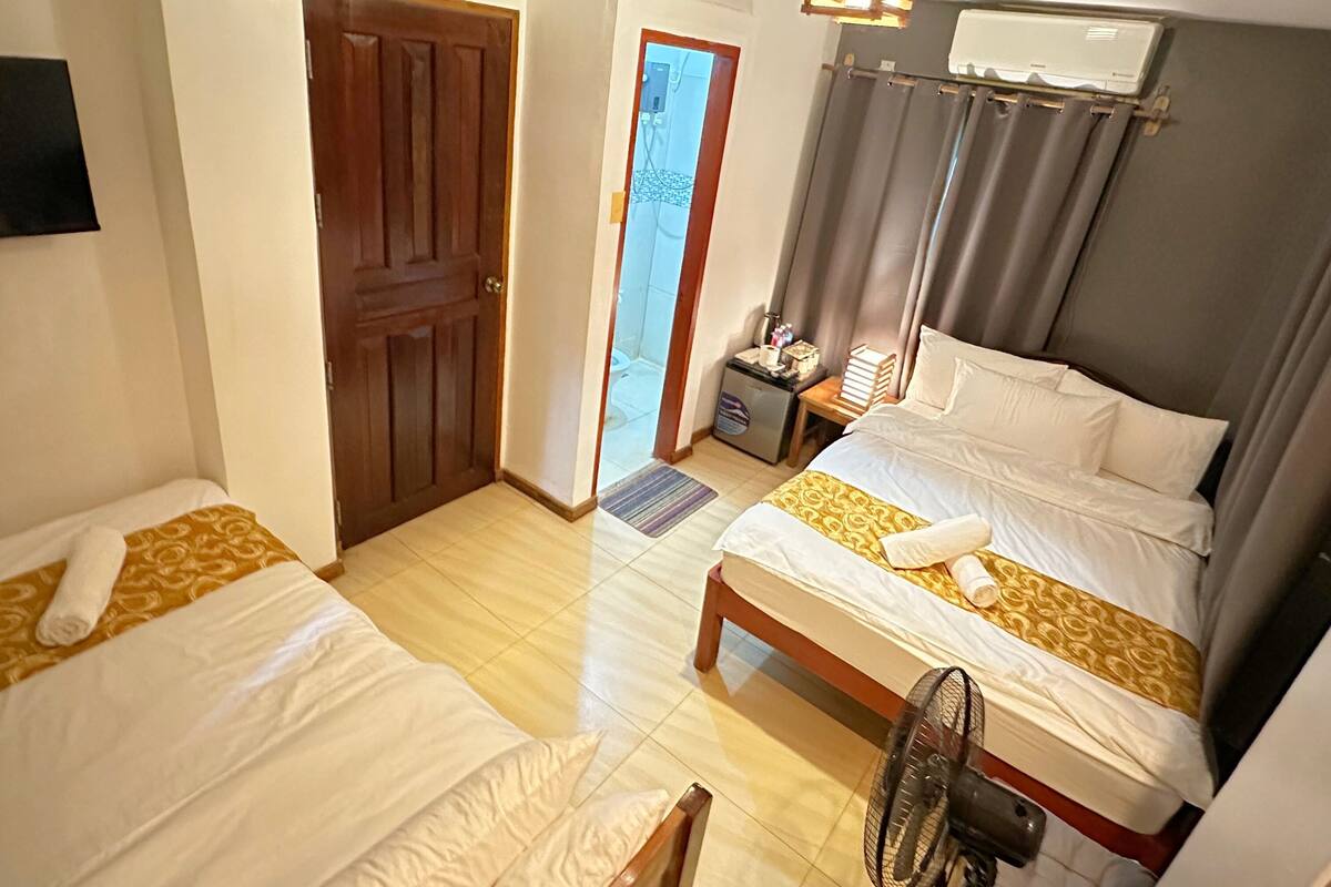 Une chambre d’hôtel avec deux lits, une porte en bois, un ventilateur de plafond et une fenêtre avec des rideaux.