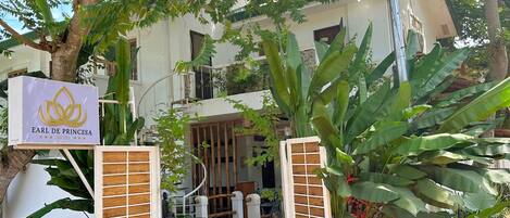 A hotel entrance with a signboard, a green carpeted path, and lush greenery.