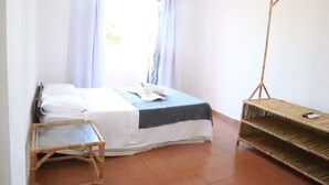 A room with a bed, a small table, a shelf, and a window with curtains. - Pousada Lisamar (Mangaratiba)