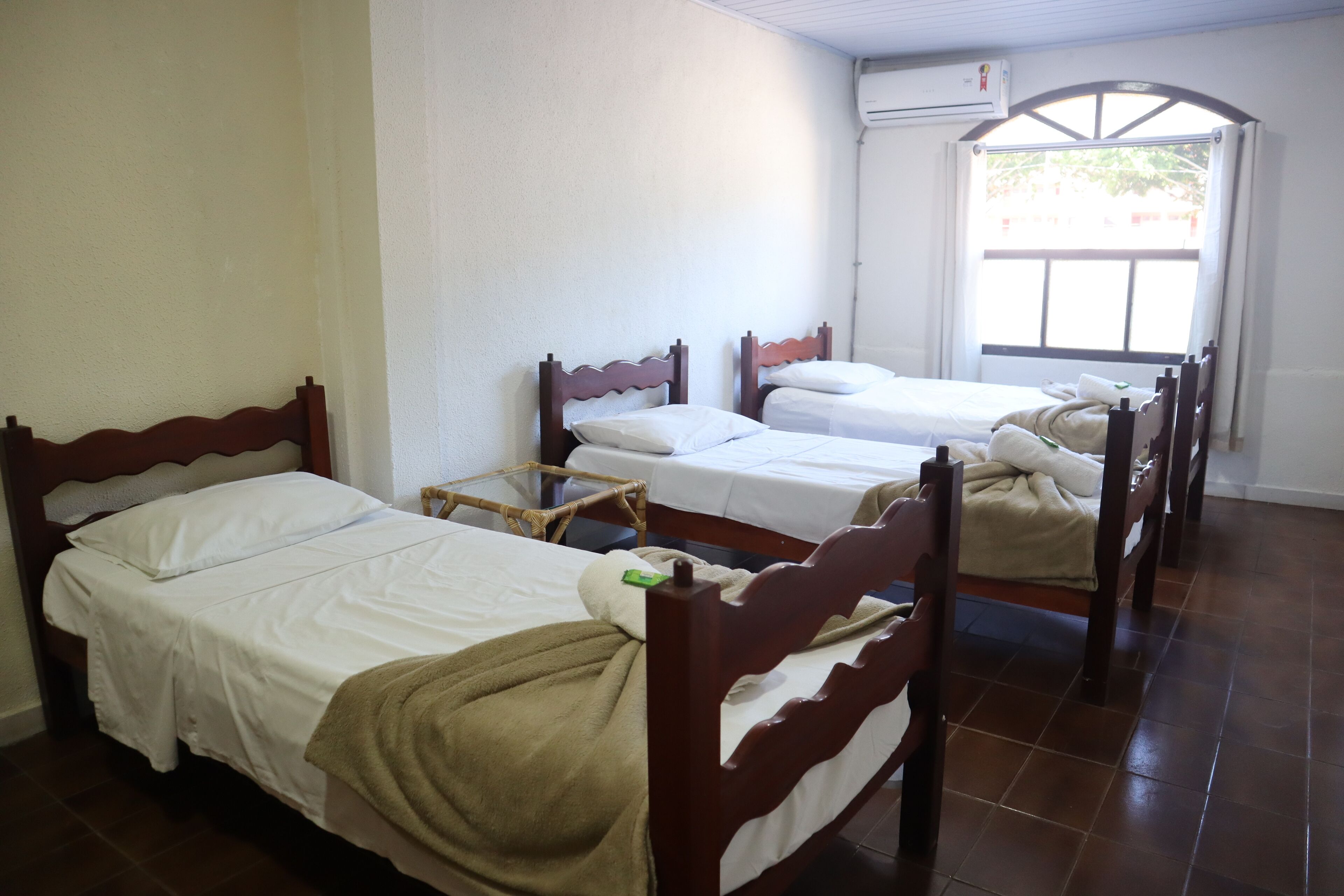 A room with four wooden beds arranged in a row, white bedding, and a window with a view of greenery outside.