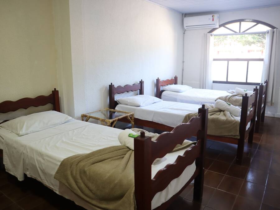 A room with four wooden beds arranged in a row, white bedding, and a window with a view of greenery outside.