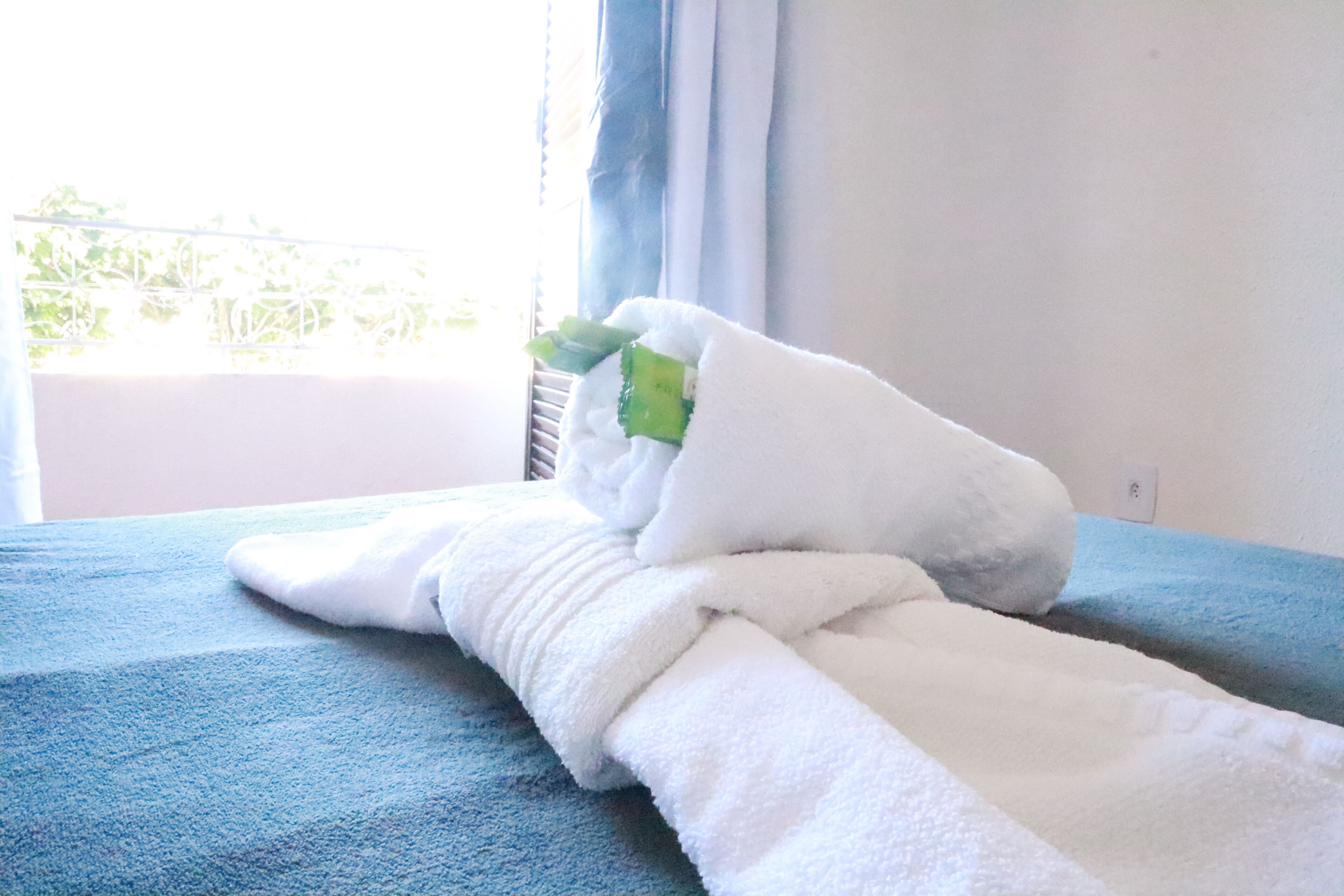 Une serviette blanche, soigneusement pliée, est posée sur un tapis de bain bleu, avec une fenêtre en arrière-plan.