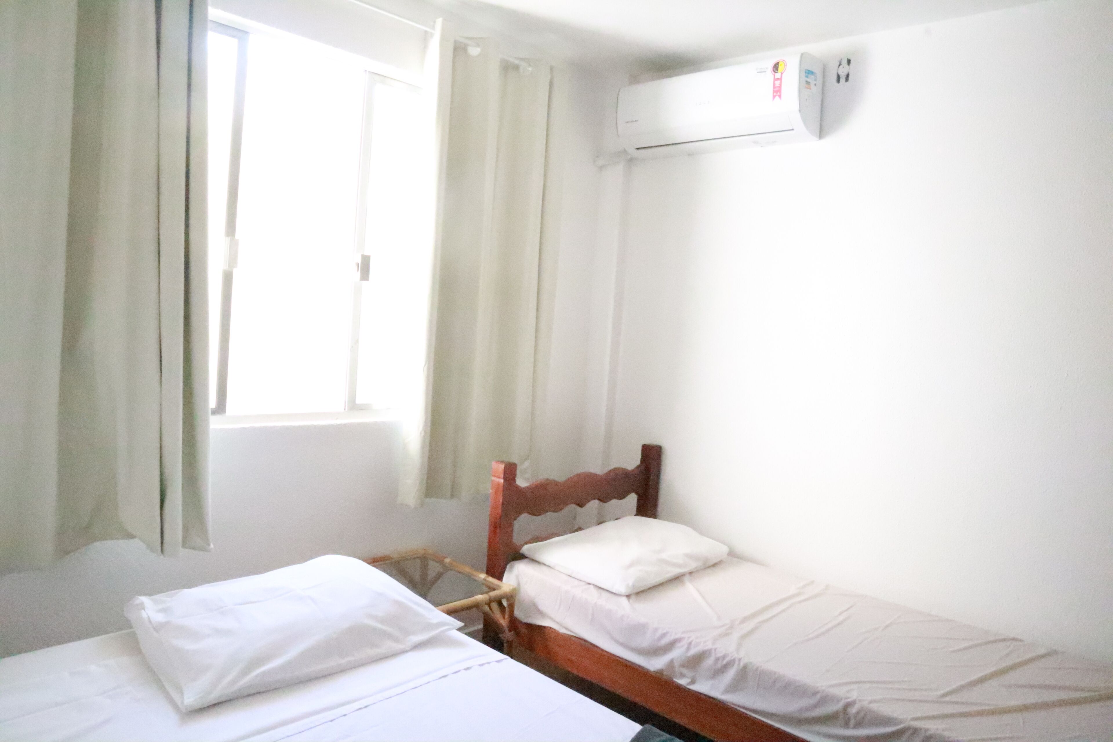 Ein Einzelbett mit Holz-Kopfteil, ein Fenster mit Vorhängen und eine Klimaanlage an der Wand.