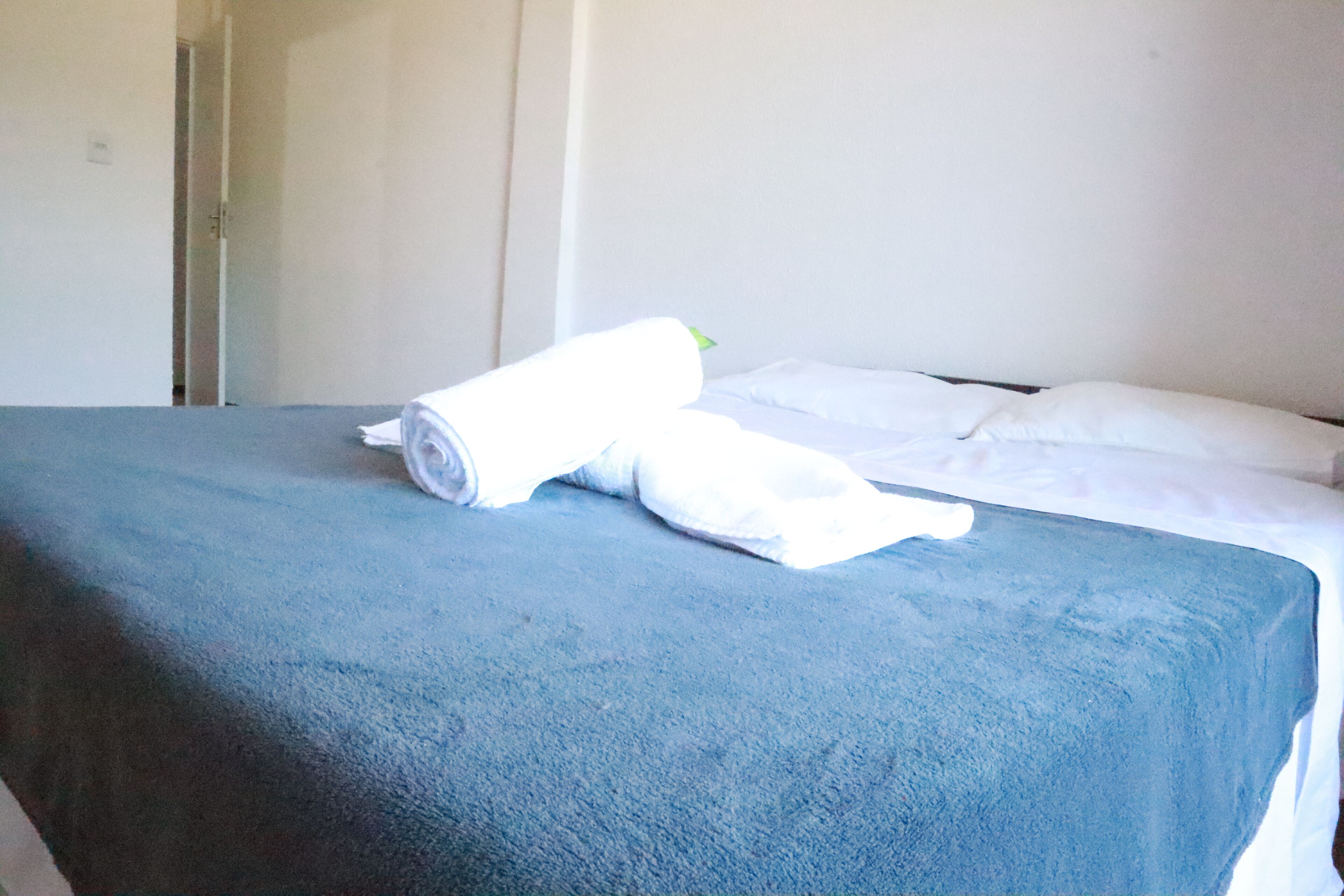 Una cama con colcha azul y almohadas blancas, una toalla blanca enrollada y una puerta entreabierta al fondo.