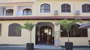 A building with a yellow facade, multiple windows with shutters, and a tiled roof. There are potted plants in front of the entrance. - Pousada Lisamar (Mangaratiba)