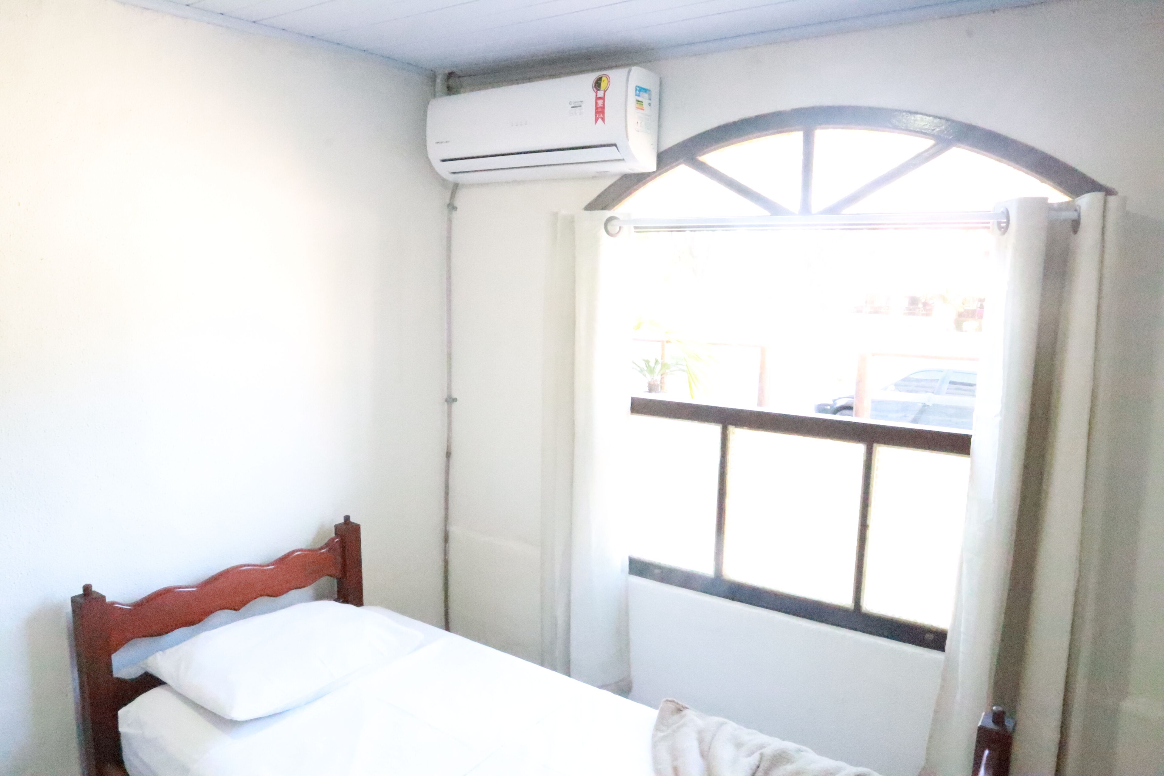 Una habitación pequeña con cama individual, cabecera de madera, ventana con reja de metal y un equipo de aire acondicionado en la pared.