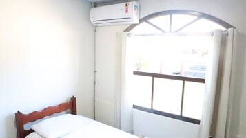Una habitación pequeña con cama individual, cabecera de madera, ventana con reja de metal y un equipo de aire acondicionado en la pared.