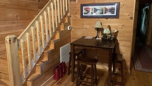 A wooden interior with a staircase, a table with chairs, a lamp, and a framed photo on the wall.