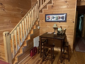 Un interior de madera con una escalera, una mesa con sillas, una lámpara y una foto enmarcada en la pared.