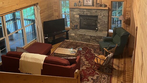 A cozy cabin interior with a stone fireplace, wooden furniture, and a large window overlooking a deck.