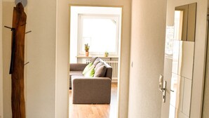 Ein Flur mit einem Holz-Kleiderständer, einer Tür und Blick in einen Wohnbereich mit einer Couch und einem Fenster.
