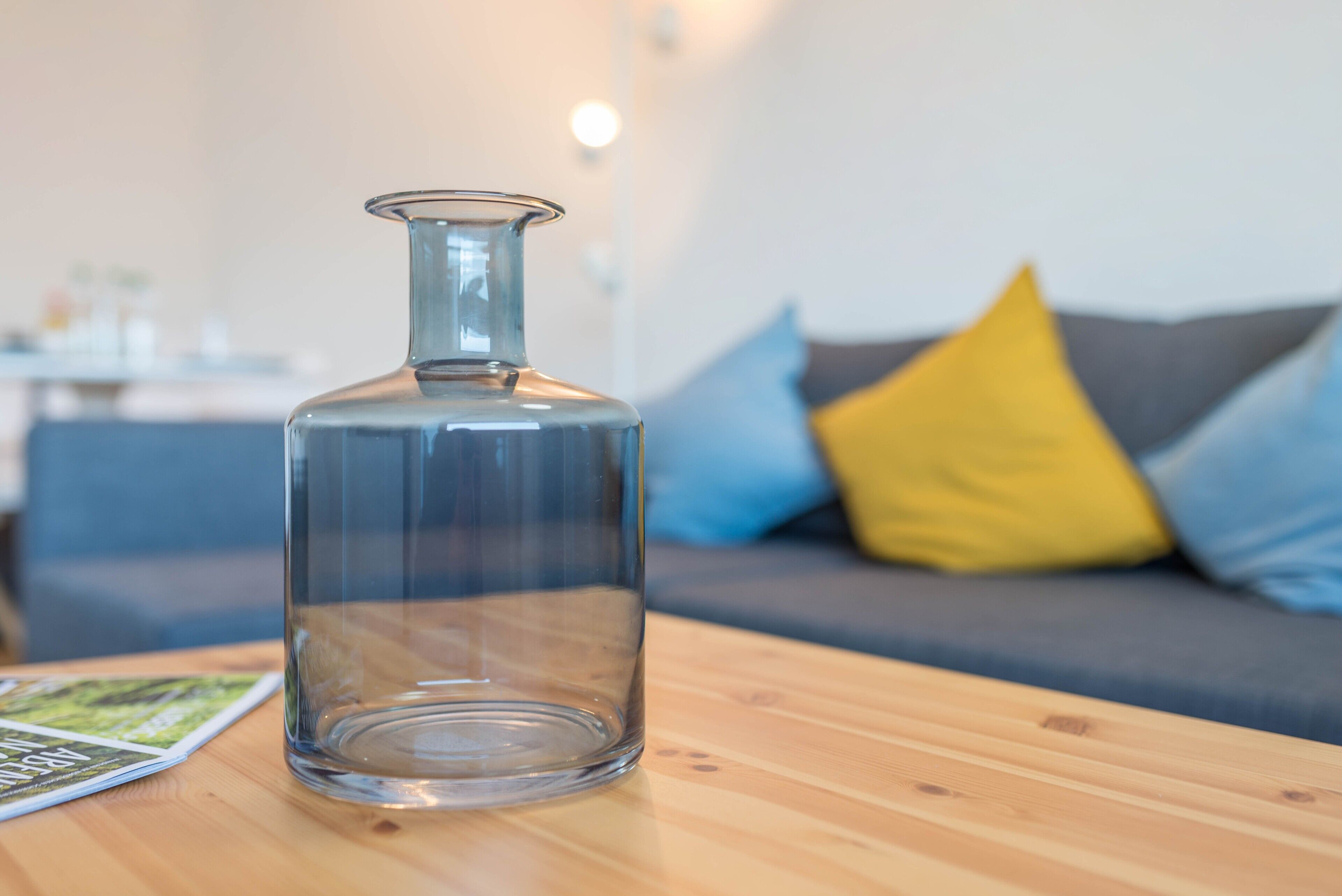 Una botella de vidrio transparente sobre una mesa de madera, con una revista, y un sofá con cojines al fondo.