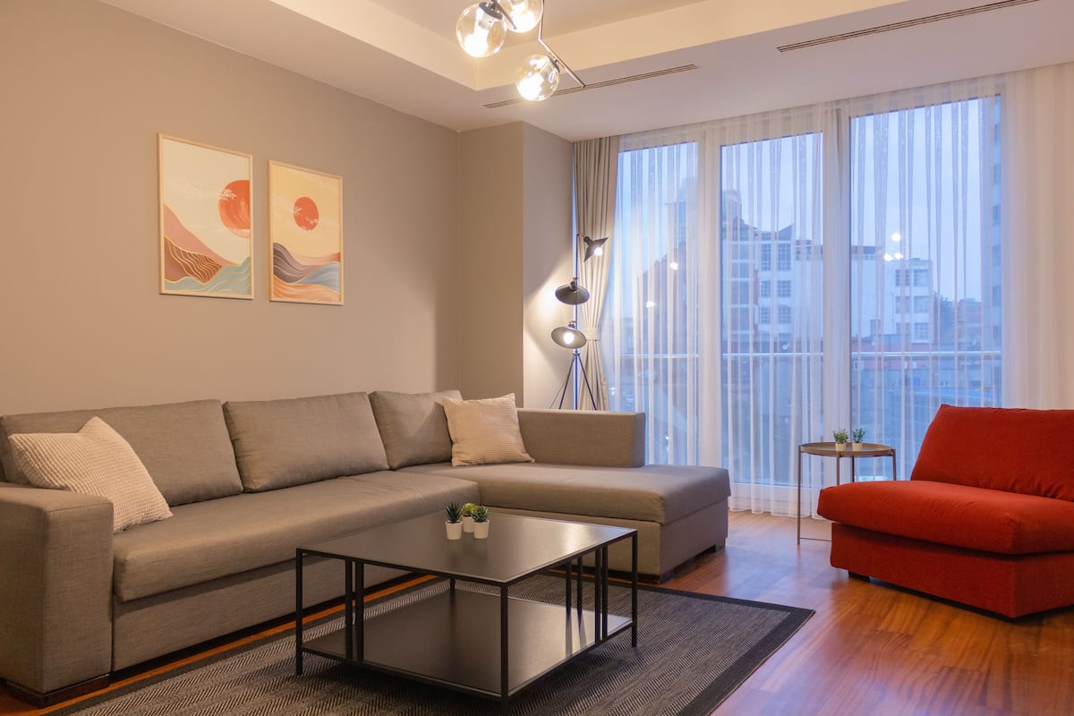 A modern living room with a grey sofa, a red armchair, a glass coffee table, and a large window with curtains.