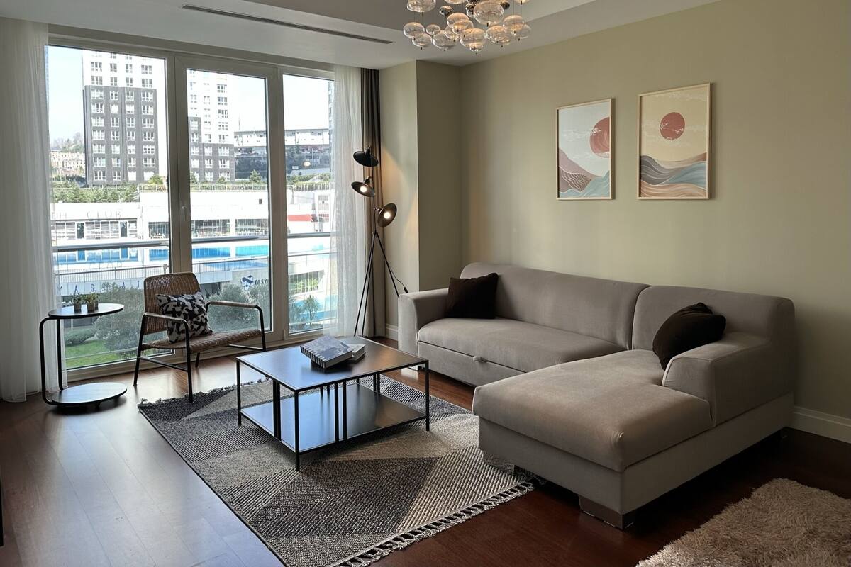 A modern living room with a sofa, a coffee table, and a chair. There is a large window with a view of buildings outside.