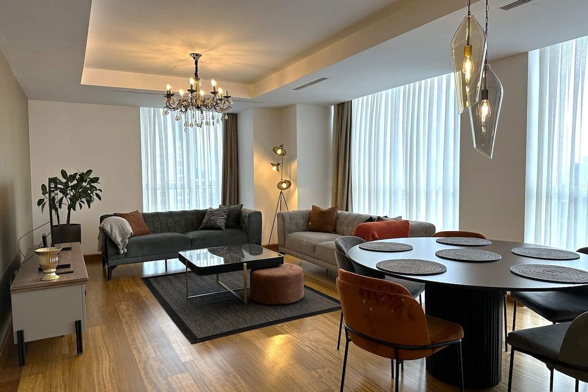 A modern living room with a sofa, coffee table, dining table, and chairs. There is a chandelier, a floor lamp, and a potted plant.