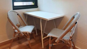 A corner room with a white desk, two wicker chairs, a window with blinds, and a framed picture on the wall.