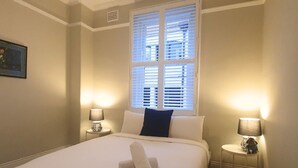 A neatly made bed with a rolled towel, two bedside lamps, a window with blinds, and a small table with a lamp and a figurine.