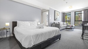 A hotel room with a large bed, a sofa, a coffee table, and a view of the city through large windows.